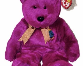 May include: A bright magenta plush teddy bear with a shiny gold ribbon tied around its neck. The bear features a black plastic nose and a small embroidered patch on its chest depicting a globe with the year 2000. A heart-shaped Ty tag is attached.