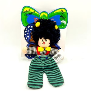 May include: A finger puppet with a black and white checkered shirt, green and black striped pants, a yellow bow tie, and a butterfly wing design. The puppet has black hair and a red nose. The packaging says "Finger Puppet" and "Try Me!"