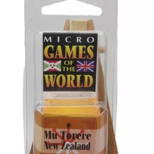 Könnte beinhalten: Ein kleiner Holzstaffelei mit einer daran befestigten gelben Box. Die Box hat ein rot-weißes Design und den Text "Mu Torere Neuseeland". Die Staffelei hat ein schwarz-weißes Etikett mit der Aufschrift "Micro Games of the World".