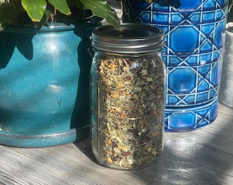 May include: A clear glass jar filled with a blend of dried herbs and flowers. The jar has a silver lid and is sitting on a wooden surface.