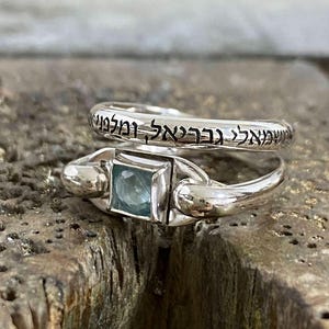 Peut inclure: Deux bagues en argent. L'une présente une pierre précieuse carrée bleu clair. L'autre bague porte une inscription en hébreu gravée sur le pourtour. Les bagues sont posées sur une surface en bois texturée.