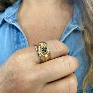 May include: A gold ring set with a black gemstone surrounded by smaller white gemstones. The ring is worn on a finger with three other gold bands.