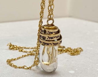 Puede incluir: Un collar de cadena de oro con un colgante de vidrio transparente. El colgante está rodeado por múltiples anillos de oro con texto hebreo inscrito en ellos.