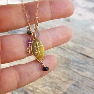 Puede incluir: Collar dorado con un colgante en forma de hoja. El colgante tiene iniciales grabadas y est&aacute; adornado con peque&ntilde;as piedras preciosas de varios colores, incluyendo verde, morado y negro. El collar es sostenido por una persona.