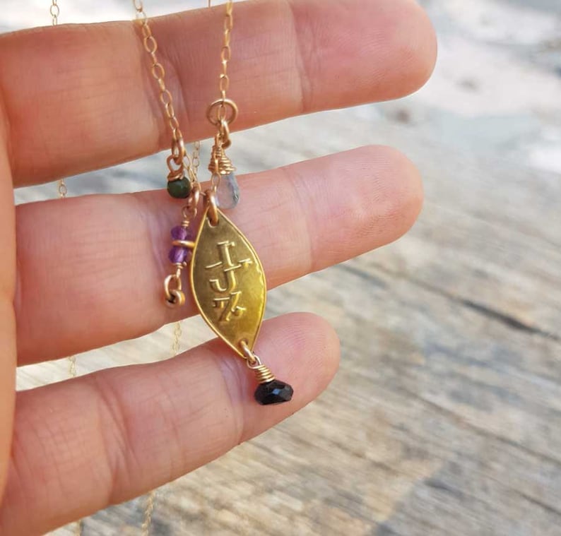Puede incluir: Collar dorado con un colgante en forma de hoja. El colgante tiene iniciales grabadas y est&aacute; adornado con peque&ntilde;as piedras preciosas de varios colores, incluyendo verde, morado y negro. El collar es sostenido por una persona.