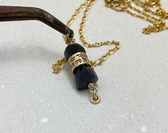 May include: A gold chain necklace with a black crystal pendant. The pendant is engraved with Hebrew text.
