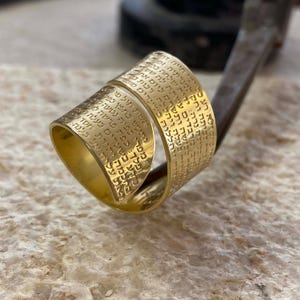 Puede incluir: Un anillo dorado con texto grabado. El anillo tiene un diseño envolvente, con el texto que cubre la superficie exterior. El anillo se muestra sobre una superficie texturizada, mostrando sus intrincados detalles y artesanía.