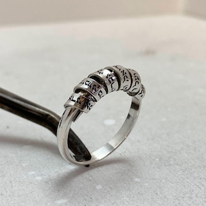 Puede incluir: Un anillo de plata con un diseño único. El anillo presenta una serie de bandas conectadas, cada una inscrita con lo que parece ser texto. El anillo se muestra sobre una superficie gris claro, mostrando sus intrincados detalles y artesanía.