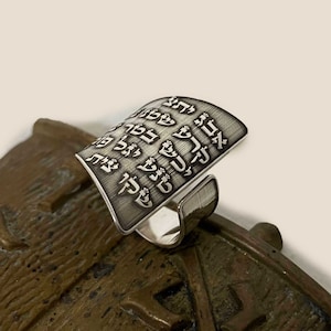 Puede incluir: Un anillo de plata con una parte superior de forma cuadrada que presenta texto en hebreo. El anillo está hecho de metal y tiene un acabado cepillado.