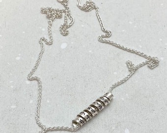 Può includere: Una collana a catena d'argento con un ciondolo composto da più anelli d'argento. Ogni anello ha un testo ebraico inciso su di esso.