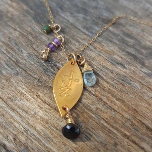 Puede incluir: Collar dorado con un colgante en forma de hoja grabado con iniciales. El collar presenta una cuenta negra, una piedra azul en forma de l&aacute;grima y peque&ntilde;as cuentas moradas y verdes. La cadena es delicada y el colgante es el punto focal.
