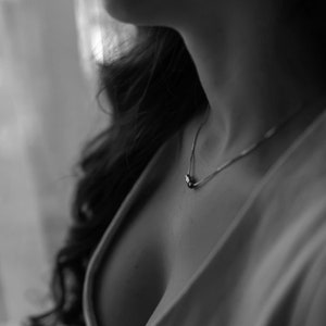 May include: A close-up of a woman's neck with a silver chain necklace with a small pendant. The woman is wearing a white shirt.