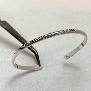 以下が含まれることがあります： 刻印されたヘブライ語のテキストが入ったシルバーのカフブレスレット。 オープンエンドのブレスレットは、研磨された仕上げと湾曲したデザインです。 テキストは黒色で、ブレスレットの上部に巻き付いています。