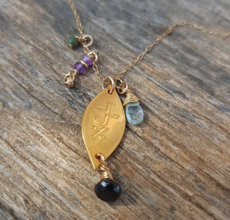 Puede incluir: Collar dorado con un colgante en forma de hoja con iniciales grabadas. Incluye una cuenta negra, una gema azul y peque&ntilde;as cuentas moradas y verdes. El collar se muestra sobre una superficie de madera.