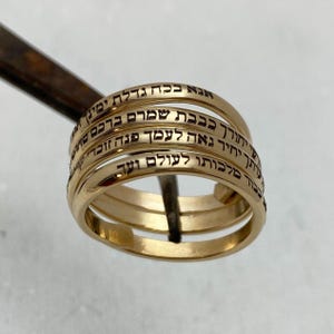 May include: A gold-colored ring with multiple bands, each engraved with Hebrew text in black. The ring is held by a metal tool against a light background. The ring's design is intricate and detailed.
