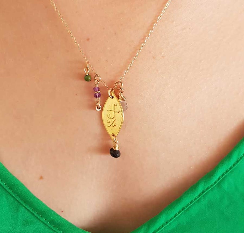 Puede incluir: Collar dorado con un colgante en forma de hoja y peque&ntilde;as cuentas de colores. El colgante tiene detalles grabados y una cuenta oscura en la parte inferior. El collar se lleva sobre una blusa verde.