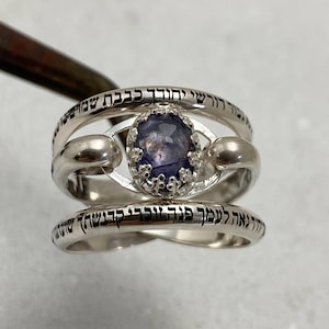 Puede incluir: Un anillo de plata con una piedra preciosa púrpura engastada en un engaste decorativo. El anillo tiene dos bandas con texto hebreo grabado. El texto dice "Shema Yisrael Adonai Eloheinu Adonai Echad", que se traduce como "Escucha, Israel, el Señor es nuestro Dios, el Señor es Uno."