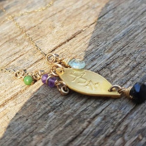 Puede incluir: Collar dorado con un colgante en forma de hoja con texto grabado. El colgante est&aacute; adornado con una cuenta negra, una gema azul claro y cuentas verdes y moradas. El collar se muestra sobre una superficie de madera.