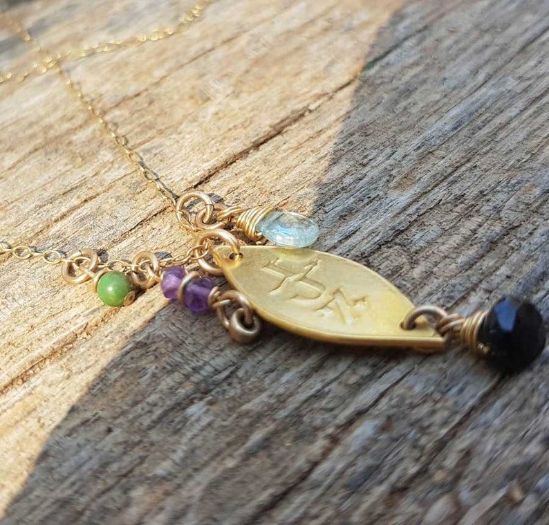 Puede incluir: Collar dorado con un colgante en forma de hoja con texto grabado. El colgante est&aacute; adornado con una cuenta negra, una gema azul claro y cuentas verdes y moradas. El collar se muestra sobre una superficie de madera.