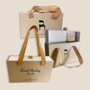 May include: Three beige gift boxes with brown ribbon handles. The boxes are decorated with the Hebrew letter 'Heh' and the text 'Ravit Hasday Jewelry' and 'www.ravithasday.com'.