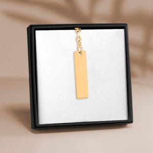 Könnte beinhalten: Ein goldfarbener, vertikaler rechteckiger Anhänger an einer zarten goldfarbenen Kette, präsentiert in einer schwarzen quadratischen Geschenkbox mit weißem Innenfutter. Der Anhänger hat eine glatte, polierte Oberfläche ohne Gravuren.
