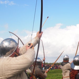 May include: A group of archers in medieval armor aiming bows at a target. The archers wear metal helmets and chainmail, with a blue sky and clouds in the background. The scene depicts a historical reenactment.