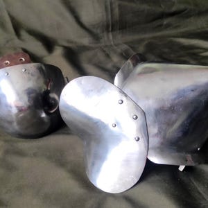 Puede incluir: Tres piezas de armadura de acero pulido, probablemente para el brazo y el hombro. Las piezas son curvas y remachadas, con una superficie reflectante. La armadura está diseñada para proteger al usuario de los golpes. Las piezas están sobre un fondo verde oscuro.