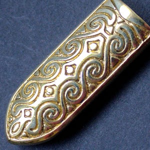 Puede incluir: Un objeto metálico dorado y alargado con un intrincado patrón de remolinos. El diseño presenta una serie de líneas curvas y formas cuadradas, creando una apariencia decorativa y ornamentada. El objeto tiene un acabado pulido, que refleja la luz.