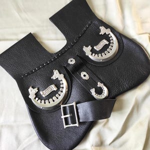 Pode incluir: Acessório de couro preto com detalhes decorativos prateados. Possui uma fivela, correias e peças metálicas decorativas com um design de rolagem. O couro tem uma aparência texturizada.