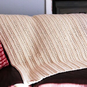 May include: A white crocheted throw blanket with a textured pattern, draped over a brown couch with a red patterned pillow.
