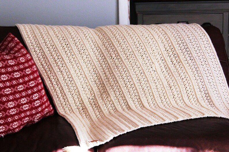 May include: A white crocheted throw blanket with a textured pattern, draped over a brown couch with a red patterned pillow.