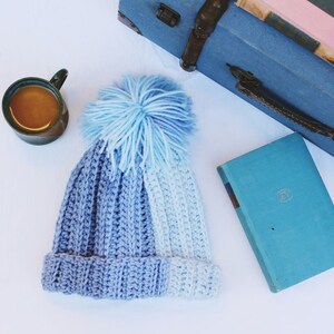 May include: A handmade, two-tone blue crochet beanie with a fluffy pom-pom. The hat is a mix of light and dark blue yarn, with a ribbed texture. The pom-pom is a lighter shade of blue. The beanie is displayed with a mug of coffee, a book, and a blue suitcase.