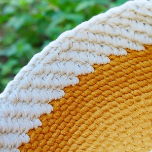 May include: A close-up of a woven macrame wall hanging with a white and yellow color scheme. The macrame is made with thick rope and has a circular shape.