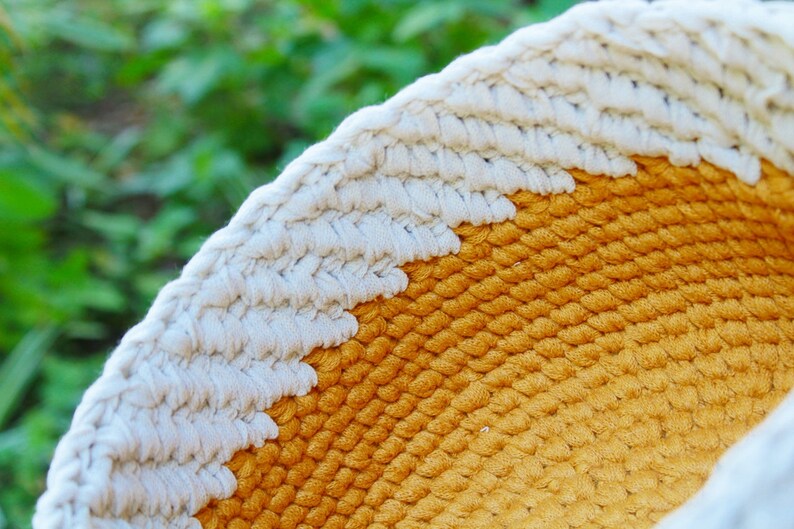 May include: A close-up of a woven macrame wall hanging with a white and yellow color scheme. The macrame is made with thick rope and has a circular shape.