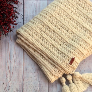 May include: A cream-colored crocheted throw blanket with a brown leather tag and white tassels.