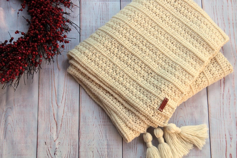May include: A cream-colored crocheted throw blanket with a brown leather tag and white tassels.