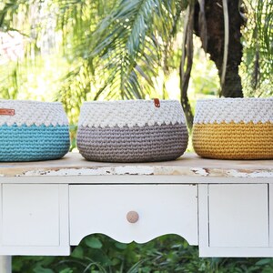 May include: Three crocheted baskets in teal, gray, and yellow with white trim. The baskets are sitting on a white table with a drawer.