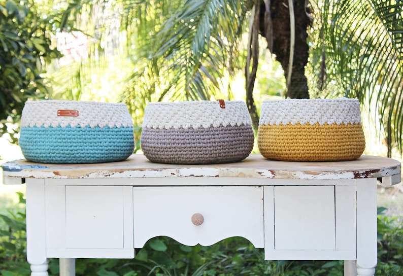May include: Three crocheted baskets in teal, gray, and yellow with white trim. The baskets are sitting on a white table with a drawer.