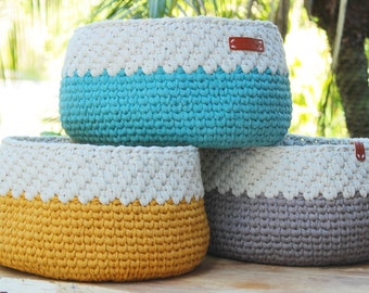 May include: Three crocheted baskets in different colours. The top basket is turquoise, the middle basket is yellow, and the bottom basket is grey. All three baskets have a white crocheted band around the top.