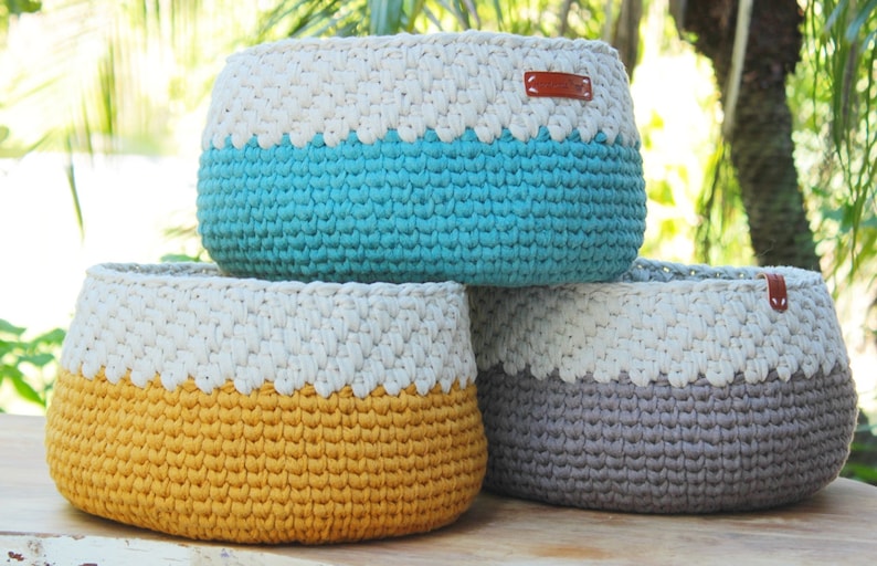 May include: Three crocheted baskets in different colors. The top basket is turquoise, the middle basket is yellow, and the bottom basket is gray. All three baskets have a white crocheted band around the top.