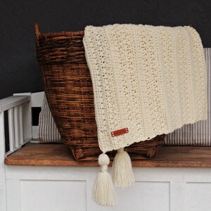 May include: A cream-colored crocheted throw blanket with tassels, folded over a wicker basket and resting on a wooden bench.