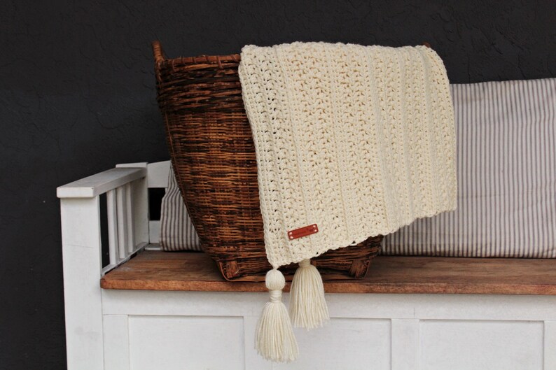 May include: A cream-colored crocheted throw blanket with tassels, folded over a wicker basket and resting on a wooden bench.