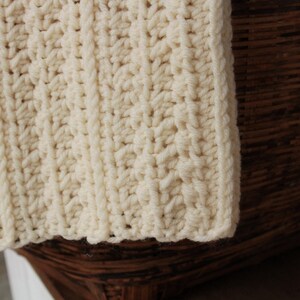 May include: A close-up of a cream-colored crocheted blanket with a ribbed texture. The blanket is draped over a brown wicker basket.