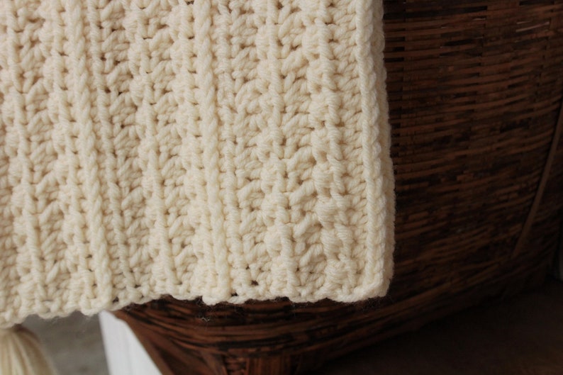 May include: A close-up of a cream-colored crocheted blanket with a ribbed texture. The blanket is draped over a brown wicker basket.