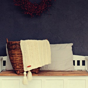May include: A white bench with a wooden top and a wicker basket with a white crocheted blanket and a striped pillow on top.