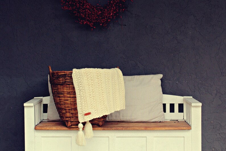 May include: A white bench with a wooden top and a wicker basket with a white crocheted blanket and a striped pillow on top.