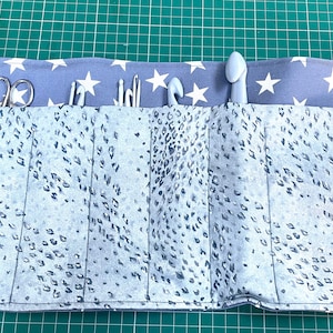 May include: A gray and white fabric roll-up case with a white star pattern. The case has six slots for holding knitting needles and crochet hooks. The case is rolled up and secured with a white ribbon.