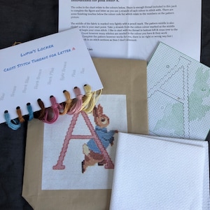 May include: Cross stitch kit for a pink letter A featuring a bunny rabbit design. The kit includes a piece of white Aida cloth, embroidery floss in various colours, and a pattern chart. The kit is labelled "Lupin's Locker Cross Stitch Threads for Letter A".