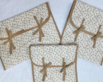 May include: A set of four quilted fabric pouches. The pouches are cream-colored with a small floral pattern and brown trim. Each pouch has a brown bow closure. The pouches are arranged on a white surface.