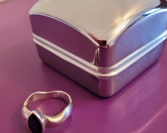 May include: A silver ring with a dark purple gemstone set in a marquise shape. The ring is sitting on a purple surface next to a silver ring box.